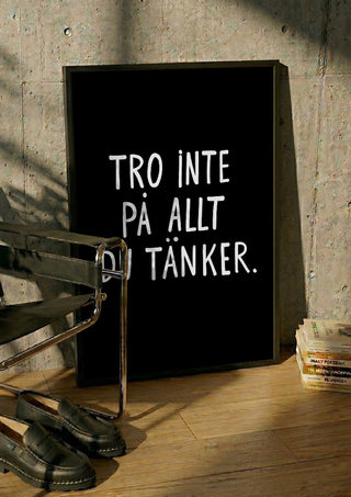 Tro inte på allt du tänker by Grafik bageri in a black frame leaning against a concrete wall by a steel chair