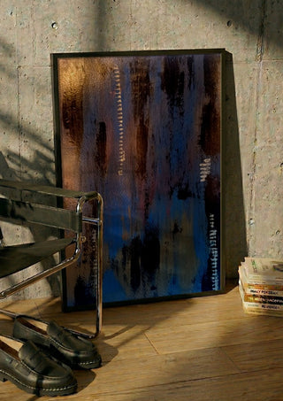 Script in Blue and Copper by Kristof ellertz in a black frame leaning against a concrete wall by a steel chair