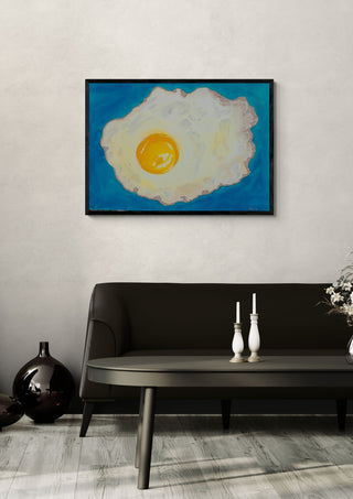 Fried egg by Sulaika Martis in a black frame on a pale wall above a dark sofa with candlesticks