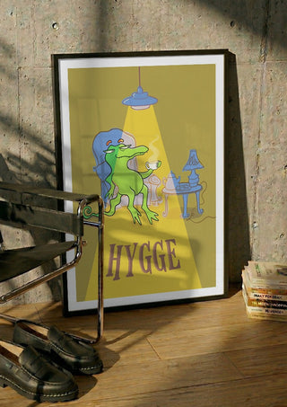 Hygge Crocodile - Yellow by Elisa Albanelli in a black frame leaning against a concrete wall by a steel chair