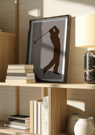 golf sport silhouette by nyai in a dark frame on a wooden shelf beside books and a lamp