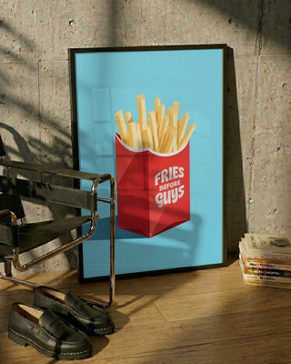 Framed print of fries in a red box labeled "Fries Before Guys" on blue background, leaning against a wall.